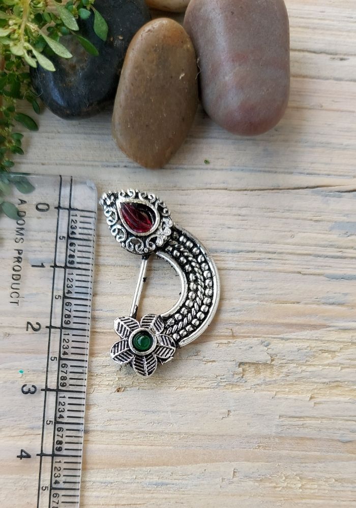 Elegant oxidised nath with intricate design featuring red and green gemstones, displayed on a wooden surface with natural stones.
