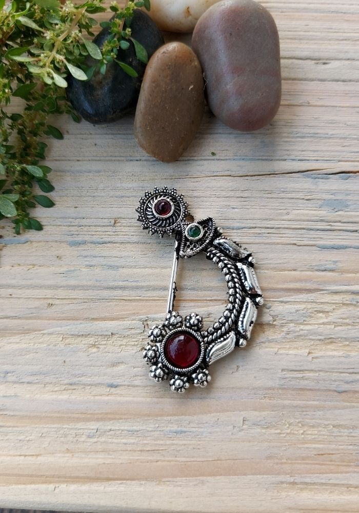 Elegant Oxidized Nath Nose Pin
Intricate floral-inspired nose pin with deep red gemstones, displayed on a rustic wooden surface amidst natural elements.