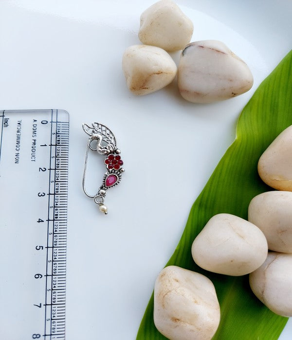 Ornate oxidized silver nath with red gemstones, displayed on a white surface alongside natural stones and a green plant leaf.