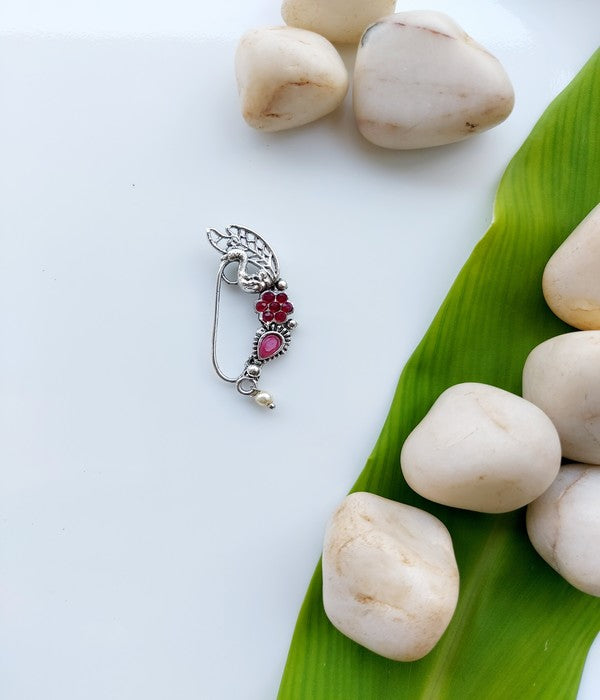 Ornate oxidized silver nath with vibrant red gem stones, surrounded by natural smooth river rocks and fresh green leaves.