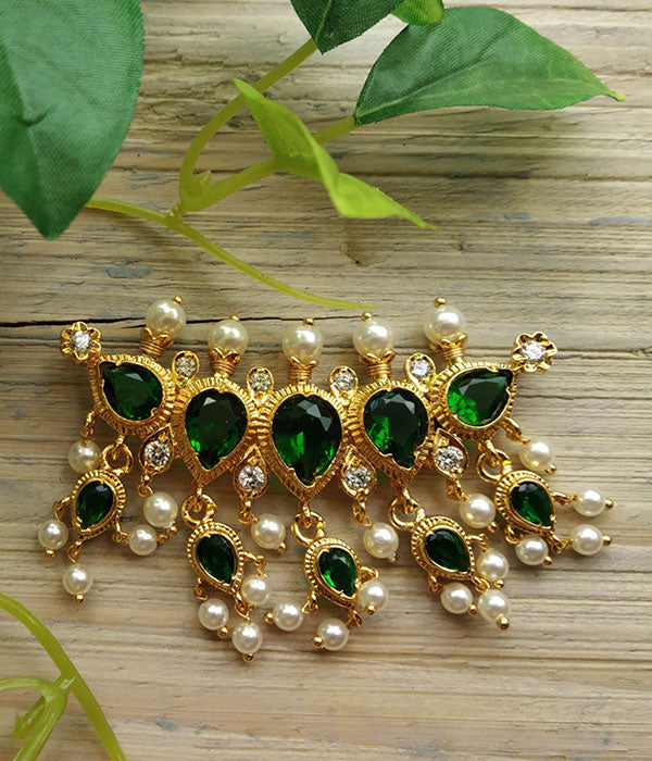 Ornate golden pendant with emerald-green gemstones and pearl accents, displayed on a rustic wooden surface with lush green leaves.