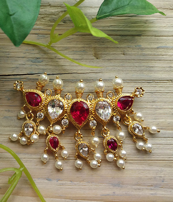 Ornate red and gold AD puneri tanmani pendant with tear-drop gemstones and dangling pearls on a weathered wooden surface with green leaves in the background.