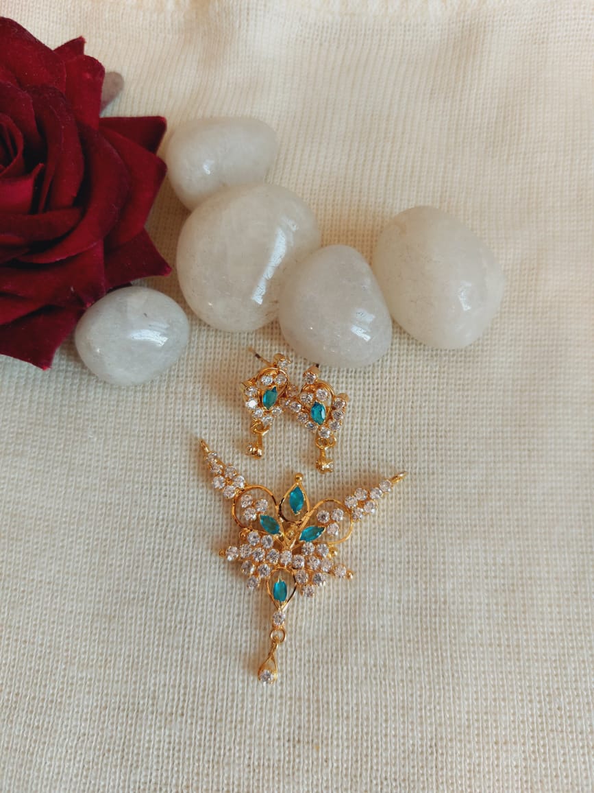 Elegant gold and gemstone pendant with vibrant blue and green accents, surrounded by white stones and a red rose on a cream fabric background.