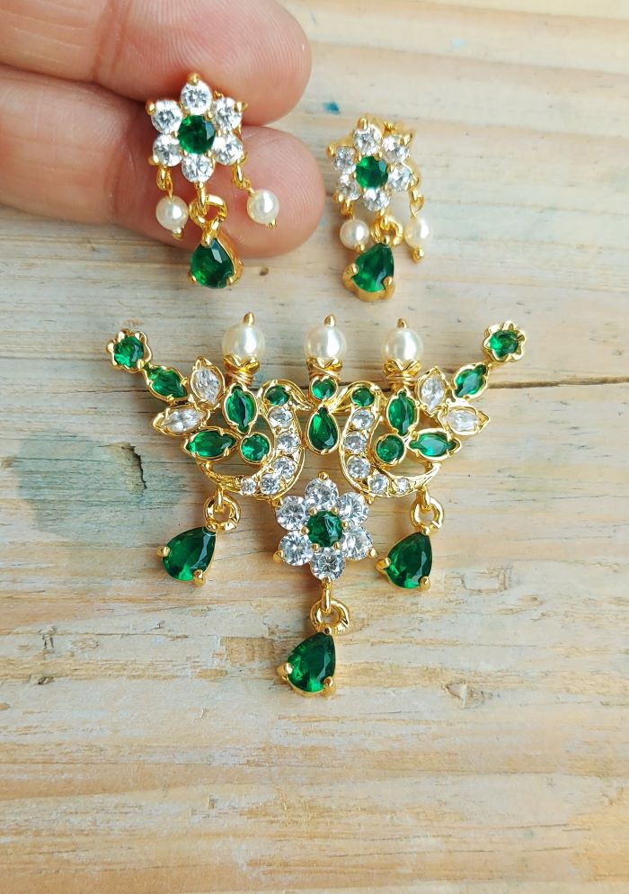 Elegant gold-toned floral designer tanmani pendant with vibrant green gemstones and sparkling white crystals displayed on a wooden surface.