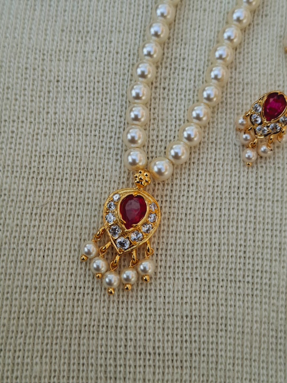 Elegant pearl necklace with red gemstone pendants, set in decorative gold-tone metal, displayed on a light knitted background.