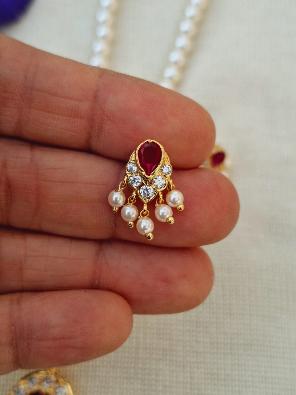 Elegant gold-toned pendant with vibrant red gemstone centered, adorned with lustrous pearls and sparkling diamonds, captured against a neutral background.