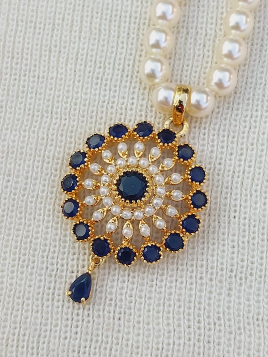 Elegant gold-tone pendant with dark blue and clear gemstones, surrounded by a pearl necklace on a light-colored knitted fabric.