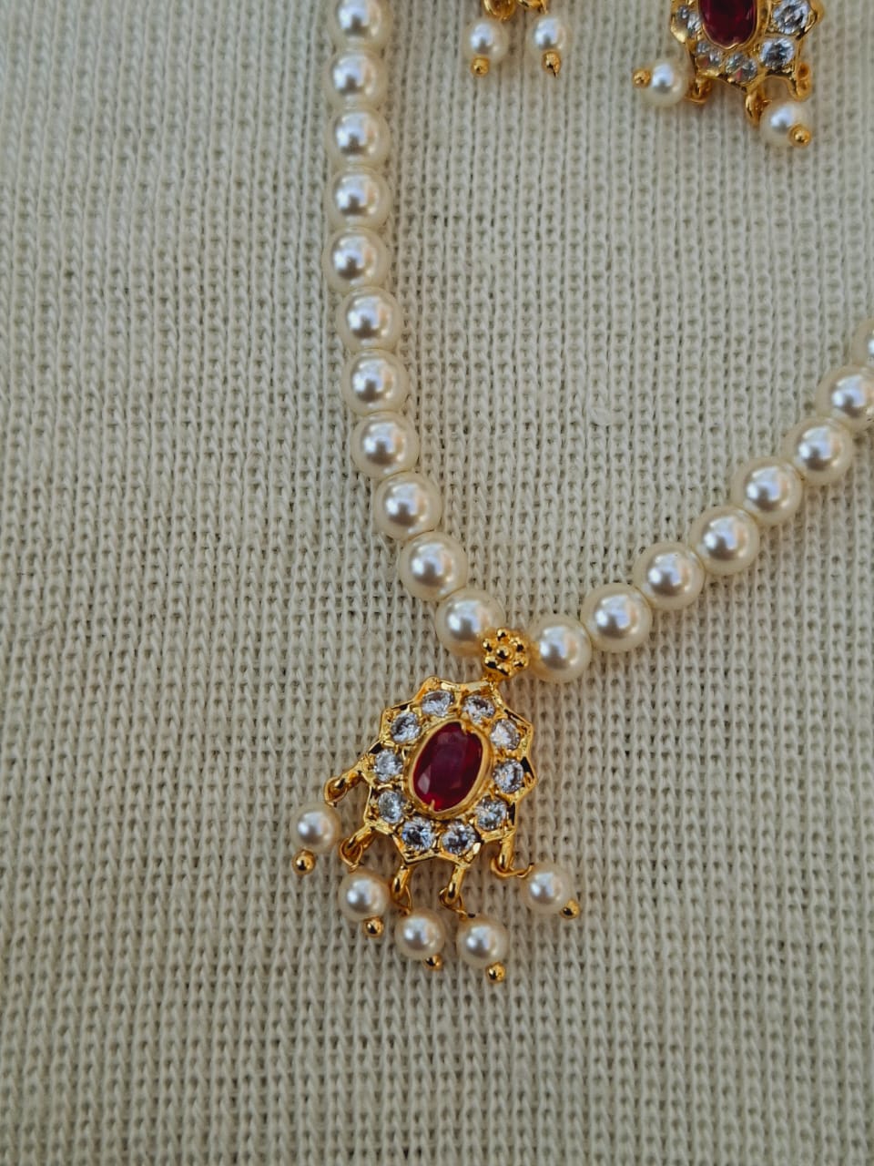Elegant pearl necklace with floral gold-toned pendant adorned with red and clear crystals, displayed on a cozy knitted gray background.