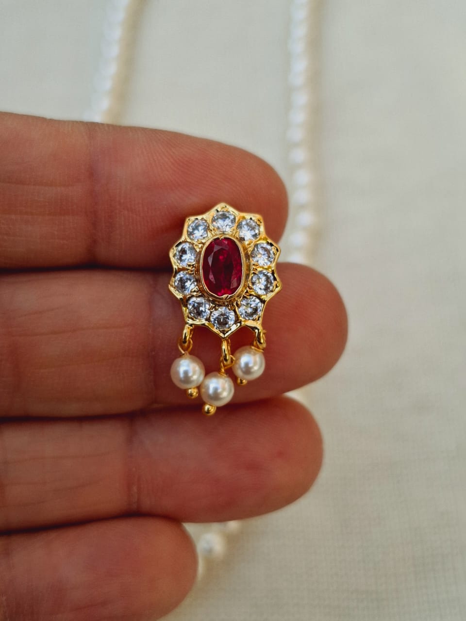 Elegant gold-toned flower-shaped pendant with a vibrant red gemstone center, surrounded by shimmering diamonds and dangling pearls, showcased on a delicate chain.