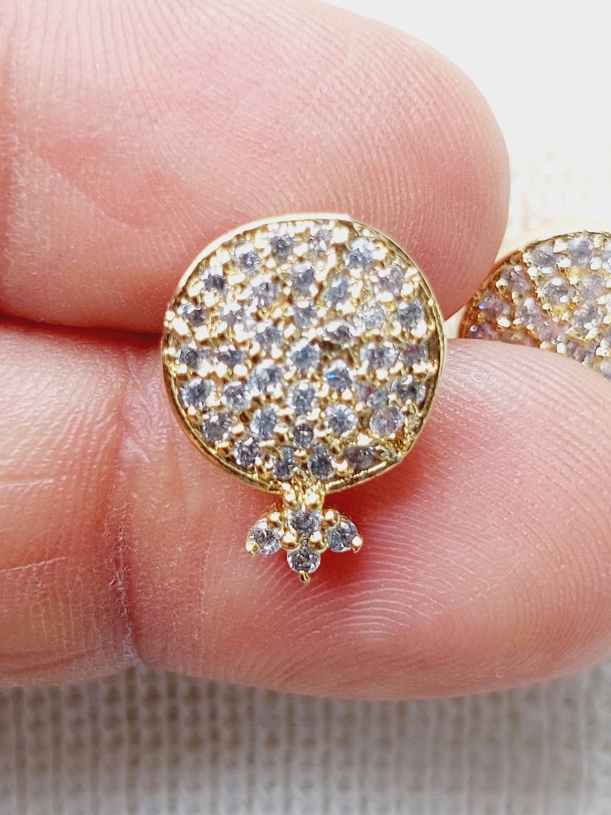 White Stone Round Earring from Mugdha Jewellery Store - Intricate golden stud earrings decorated with sparkling white stones, showcased in a close-up finger shot.