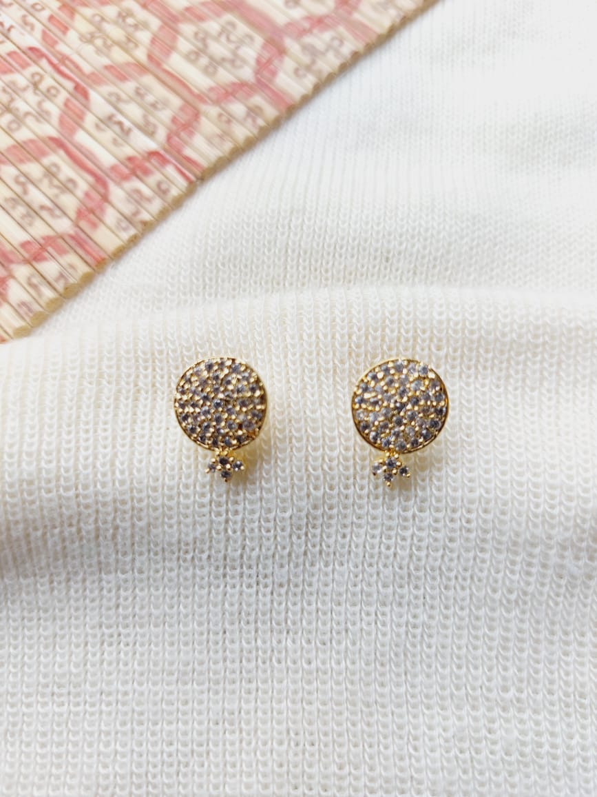 Elegant diamond-studded gold stud earrings from Mugdha Jewellery Store, placed on a soft white knitted fabric and accented by a vibrant patterned textile.