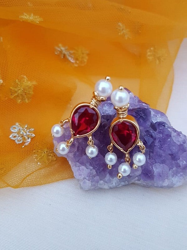 Beautiful dark pink stone tanmani earrings with pearls, placed on a vibrant purple amethyst crystal cluster against an orange background with snowflake patterns.
