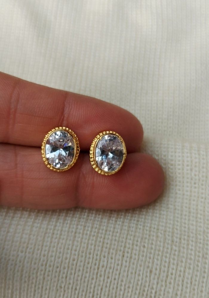 Oval-shaped white stone stud earrings with gold-toned metal frames, placed on a light-colored fabric background.