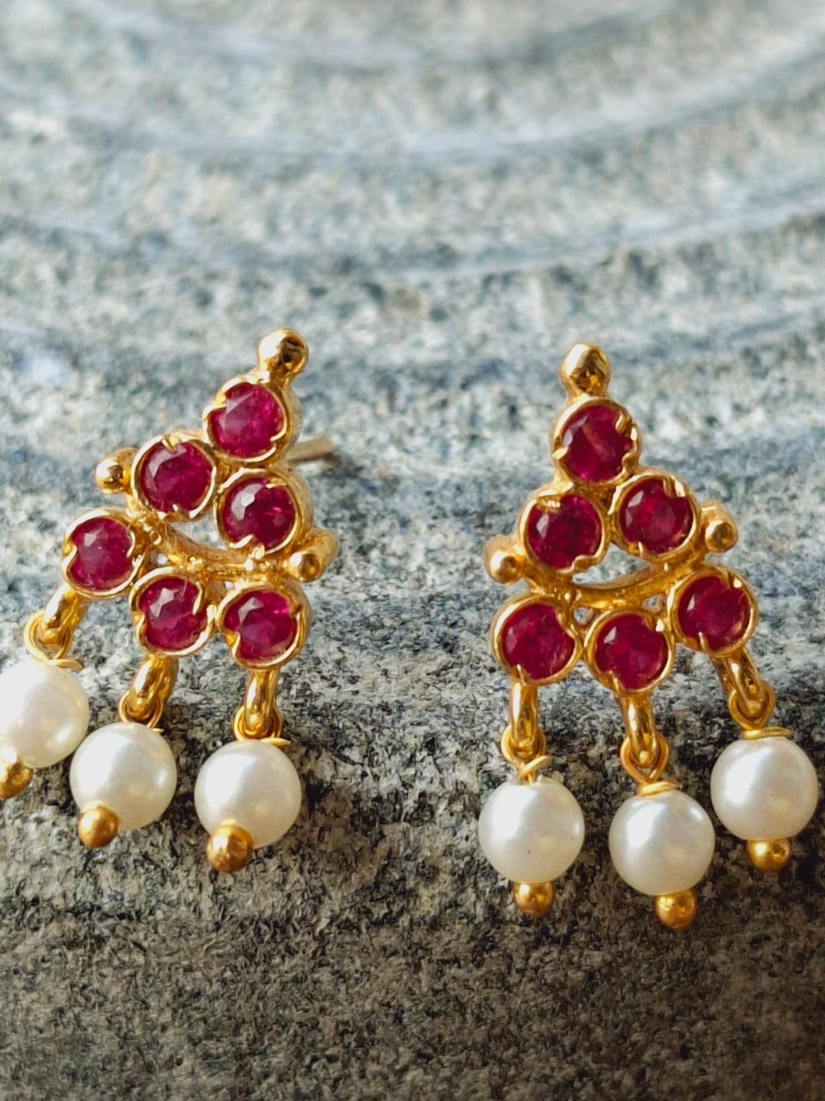 Elegant gold-toned Puneri earrings adorned with vibrant red gemstones and lustrous pearls, displayed on a textured grey background.