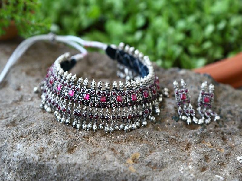 Ornate Silver and Fuchsia Jewellery Set: Detailed silver choker necklace with fuchsia gemstones and matching dangling earrings displayed on a natural stone surface against a blurred green background.