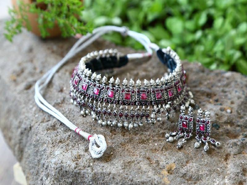 Intricate Silver Copper Mixed Laffa Necklace from Mugdha Jewellery Store
A stunning piece of jewelry featuring an ornate necklace with a mix of silver and copper accents, decorated with colorful gemstones and intricate patterns, placed on a natural stone backdrop amidst lush greenery.