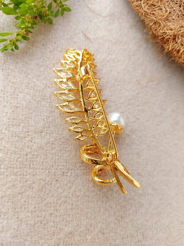 Golden leaf feather brooch with white stones on grey surface with greenery in the background.
