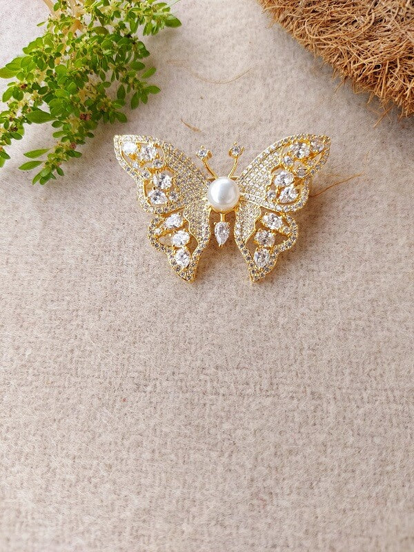 Elegant gold-colored butterfly brooch adorned with sparkling crystals and a pearl centerpiece, displayed on a textured natural surface.