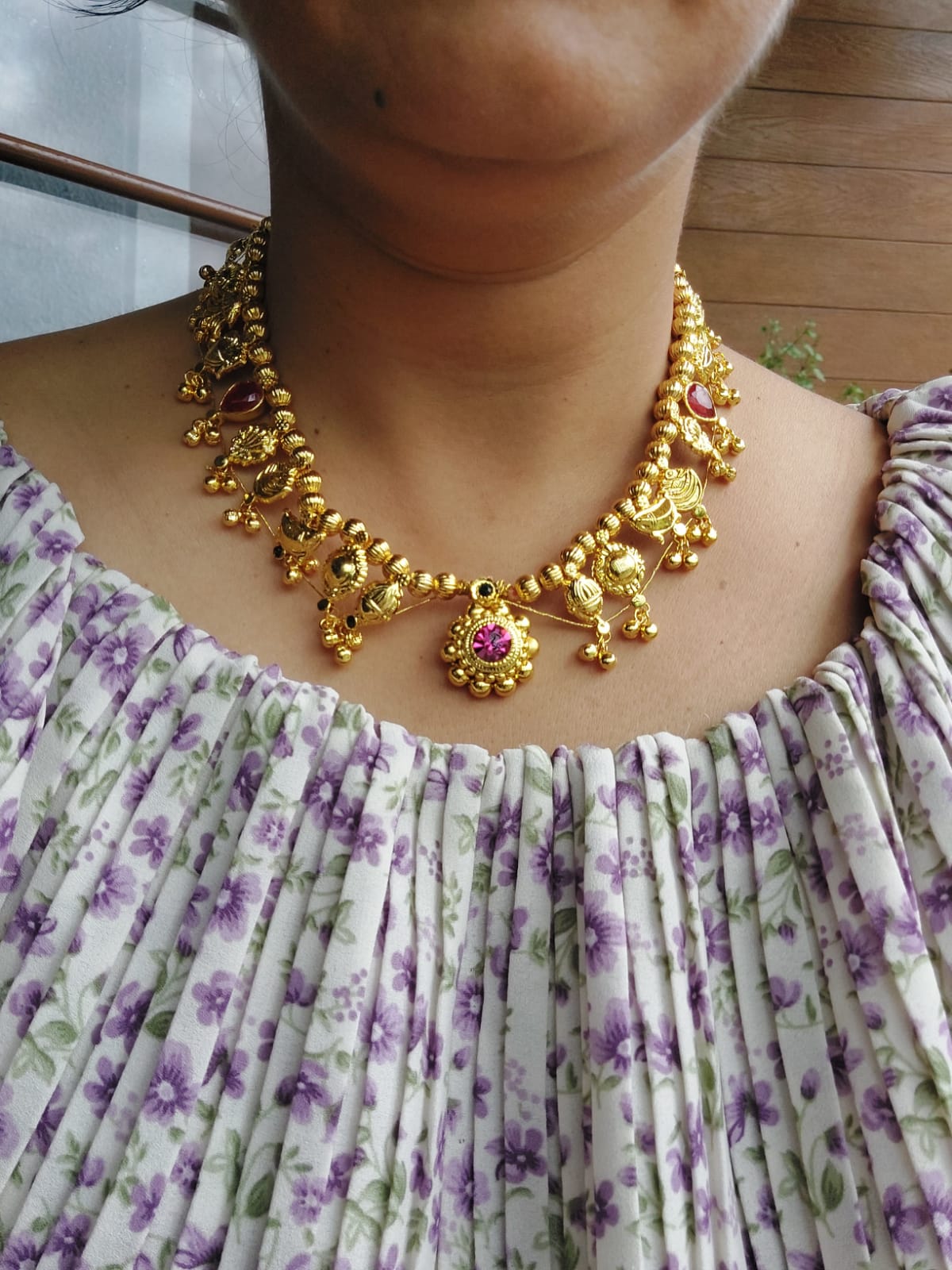 Round Bead Red Stone Kolhapuri Saaj Short, a vibrant gold-tone necklace adorned with intricate beads and a red stone centerpiece, displayed on a woman wearing a floral patterned dress.
