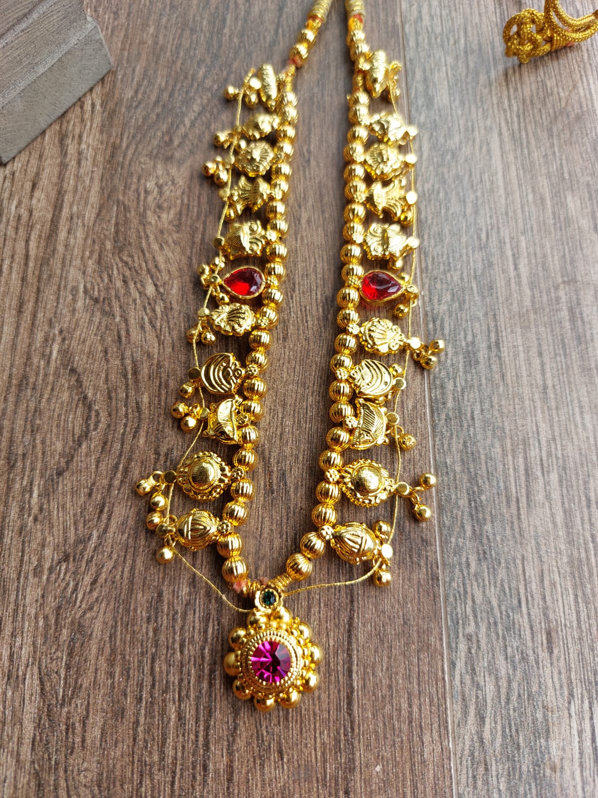 Ornate golden Kolhapuri saaj short necklace featuring round beads and intricate floral motifs with vibrant red stone accents, displayed on a wooden surface.