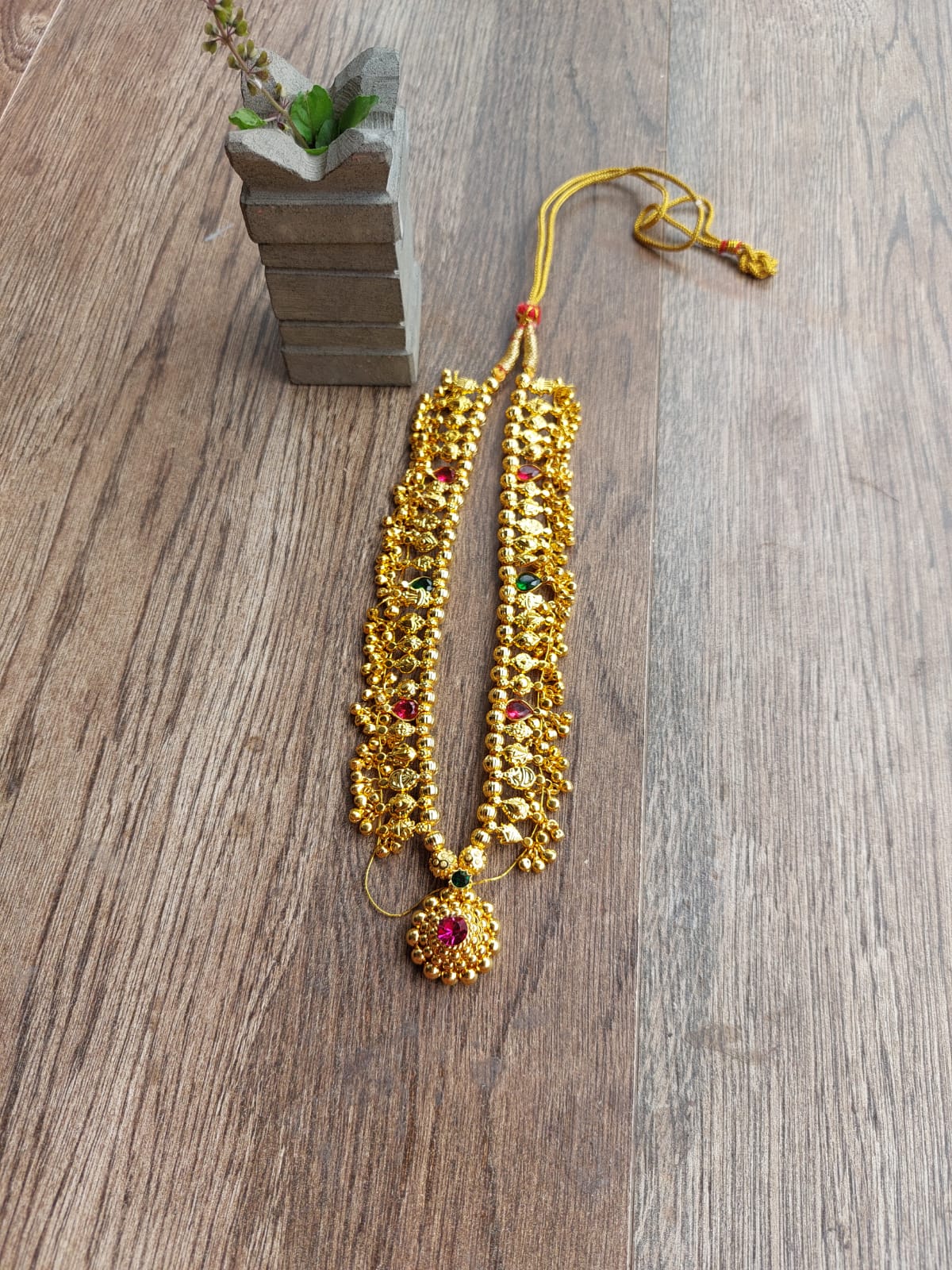 Vibrant gold-tone beaded Kolhapuri saaj short necklace with a central red gemstone accent, displayed on a wooden background.