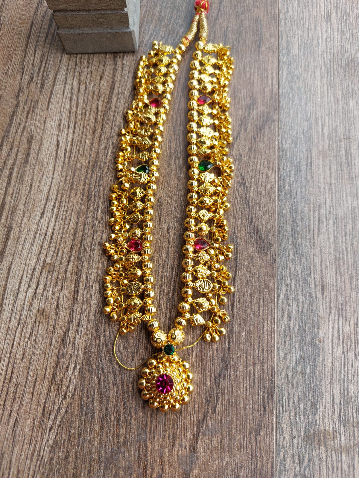 Ornate gold-tone Kolhapuri saaj necklace with multicolored gemstones and beads, displayed on a wooden surface.