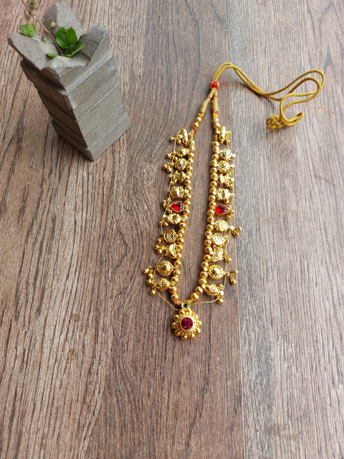 Stunning gold-toned necklace with beaded and studded design, featuring vibrant red stone accents, placed on a rustic wooden surface with a potted succulent in the background.