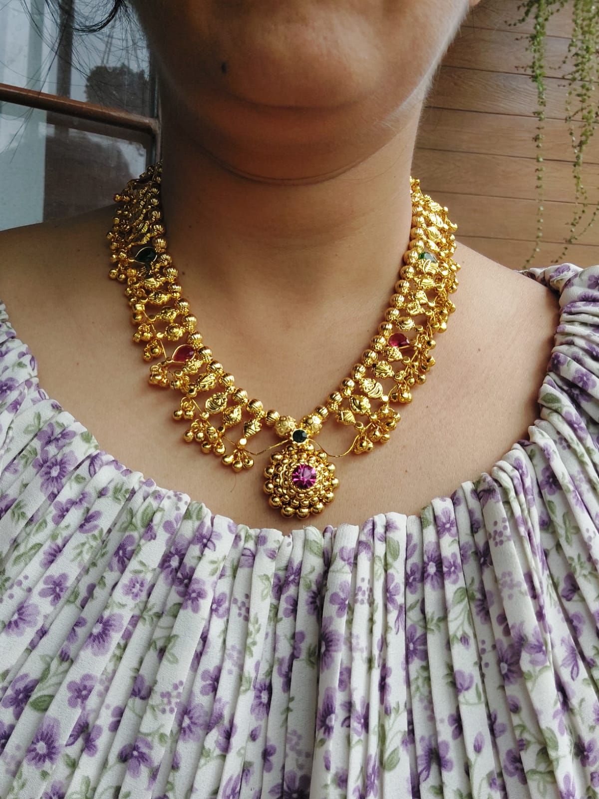Round Bead Multicoloured Heavy Kolhapuri Saaj Short, a vibrant gold-tone necklace with a central pendant, adorning the model's neck against a floral patterned dress.