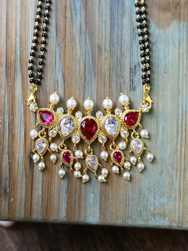 Exquisite gold-toned mangalsutra with ornate heart-shaped ruby and diamond pendants, surrounded by cascading pearl beads, displayed against a rustic wooden backdrop.
