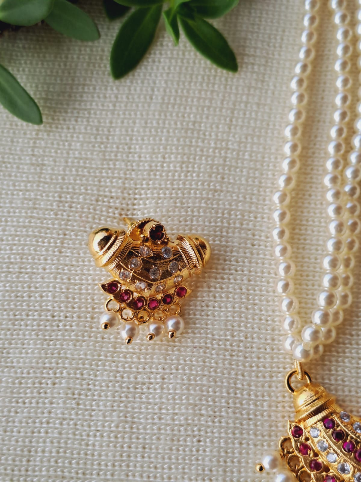 Ornamental golden pendant with textured patterns and red gemstones, displayed on a pearl necklace against a neutral background with green leaves.
