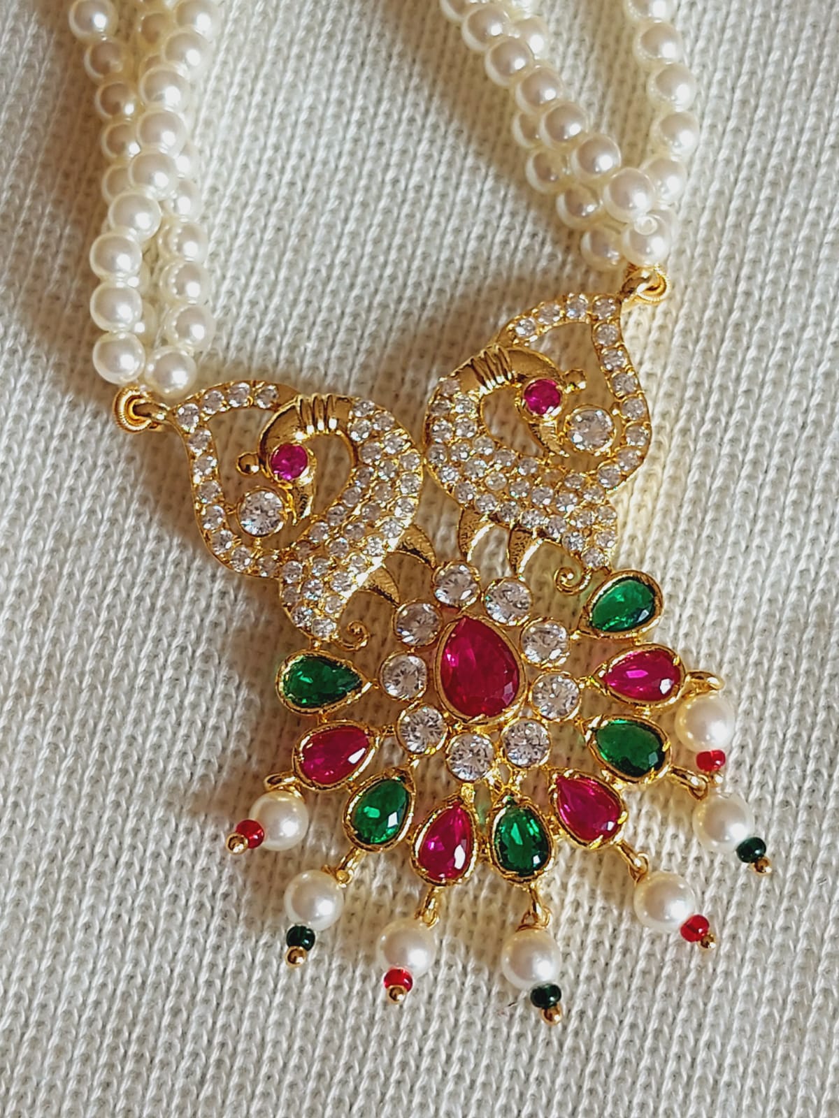 Exquisite golden peacock pendant adorned with sparkling multicolored stones and pearls, elegantly displayed on a cream-colored knitted surface.