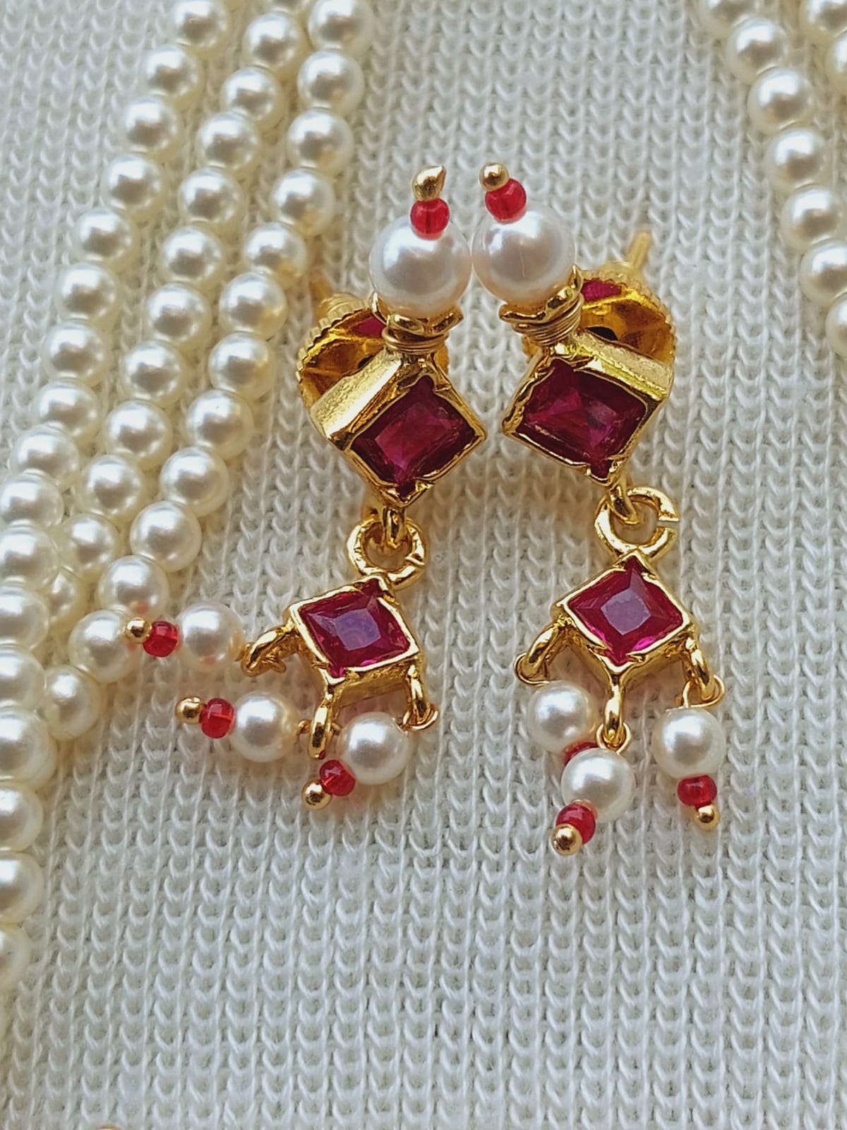Elegant pearl and ruby earrings on a white knitted background from the Mugdha Jewellery Store. The earrings feature shimmering pearls, sparkling ruby stones, and a graceful golden design.
