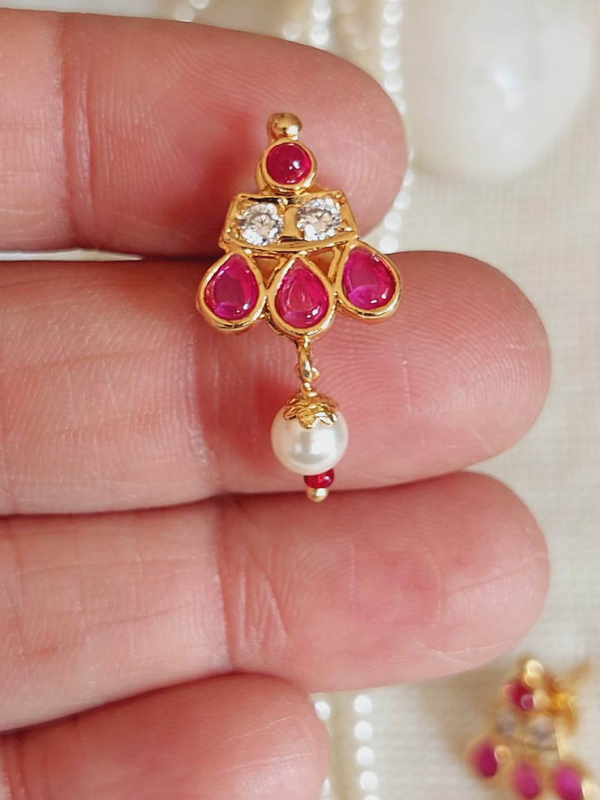 Elegant gold-toned Tanmani pendant with vibrant red gemstones and sparkling clear crystals, accented by a delicate pearl drop, showcased on a human hand.