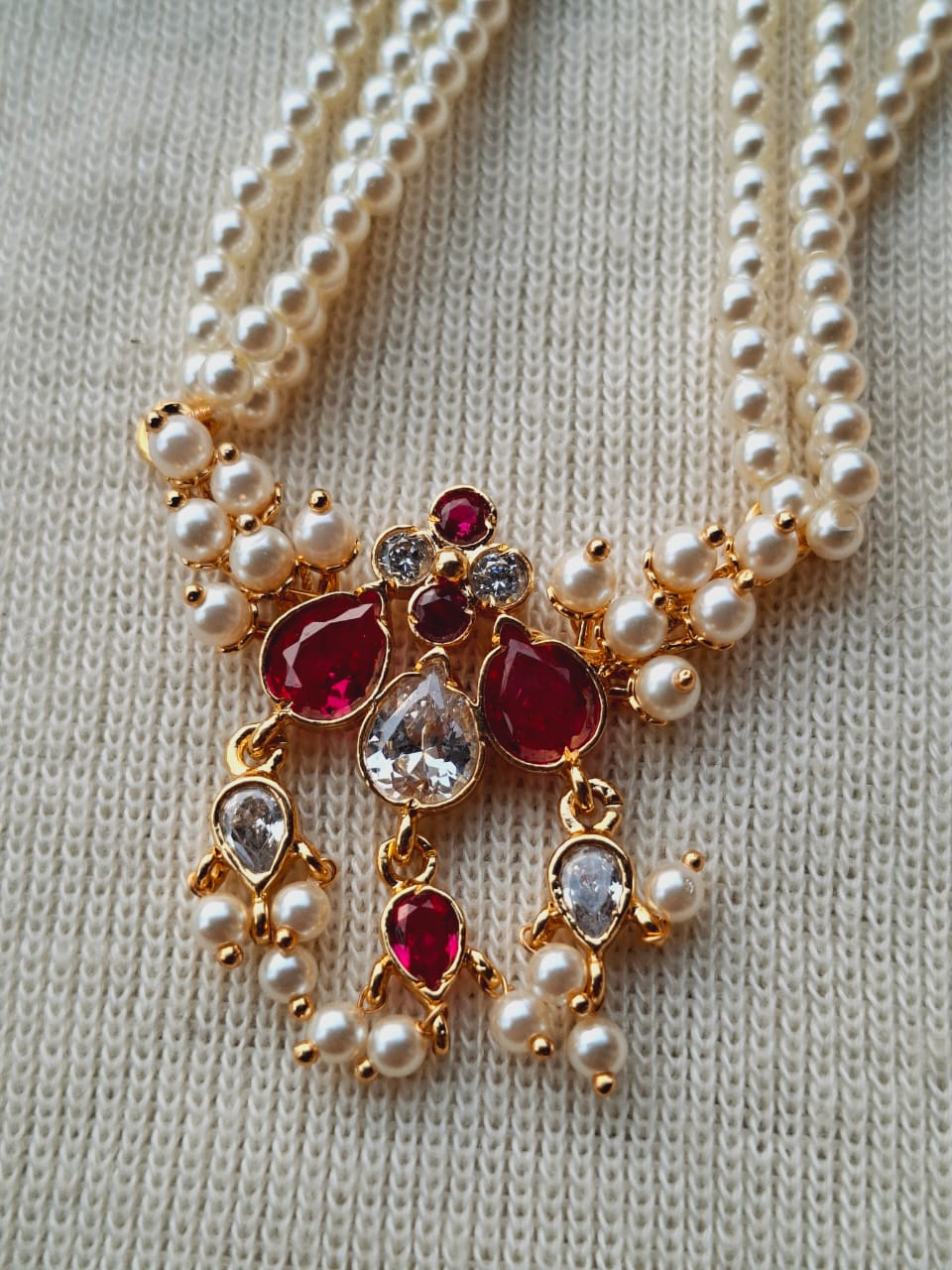 Elegant gold-toned necklace with multiple strands of lustrous pearls accented by red and clear gemstones, forming an intricate floral-inspired pendant.