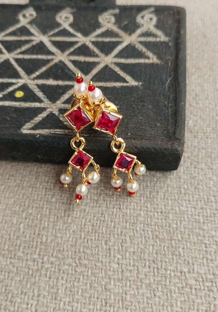 Ornate red gemstone earrings with dangling pearls, displayed on a textured dark background.