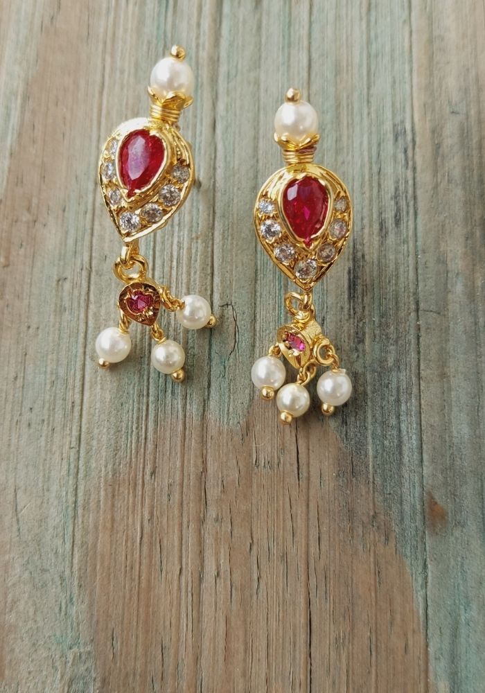 Elegant gold-plated pendant earrings featuring red and white gemstones, adorned with lustrous pearls, displayed on a rustic wooden surface from the Mugdha Jewellery Store.