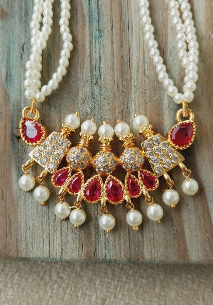 Classic Karwari Pendant Tanmani - Elegant gold-tone necklace with red gemstones, sparkling crystals, and lustrous pearls, displayed on a weathered wooden surface.