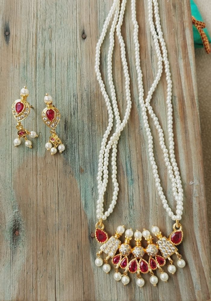 Classic multistrand pearl necklace and matching golden earrings with red gems, displayed on a rustic wooden background from Mugdha Jewellery Store.