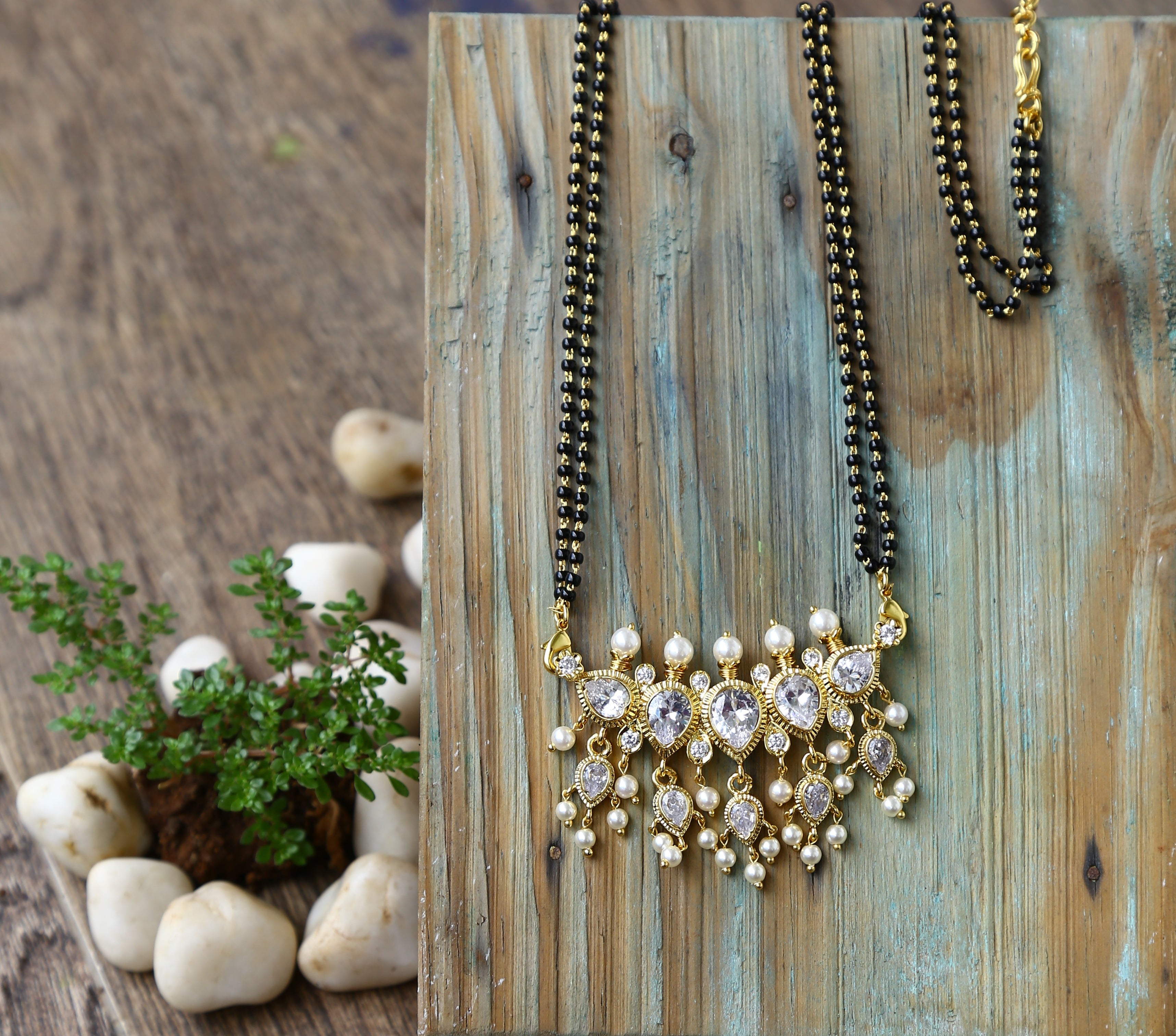 Elegant gold mangalsutra with 5 stones hanging on rustic wooden surface surrounded by natural stones and greenery.
