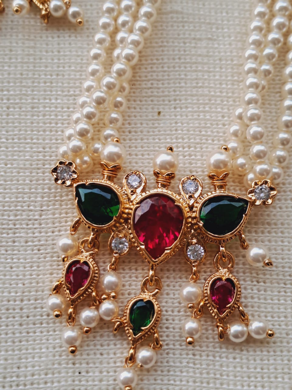 Multicoloured 3 Stone Puneri Tanmani: Ornate gold-toned pendant showcasing a large central ruby-colored gemstone surrounded by smaller green and ruby accents, adorned with shimmering pearl beads against a textured ivory background.