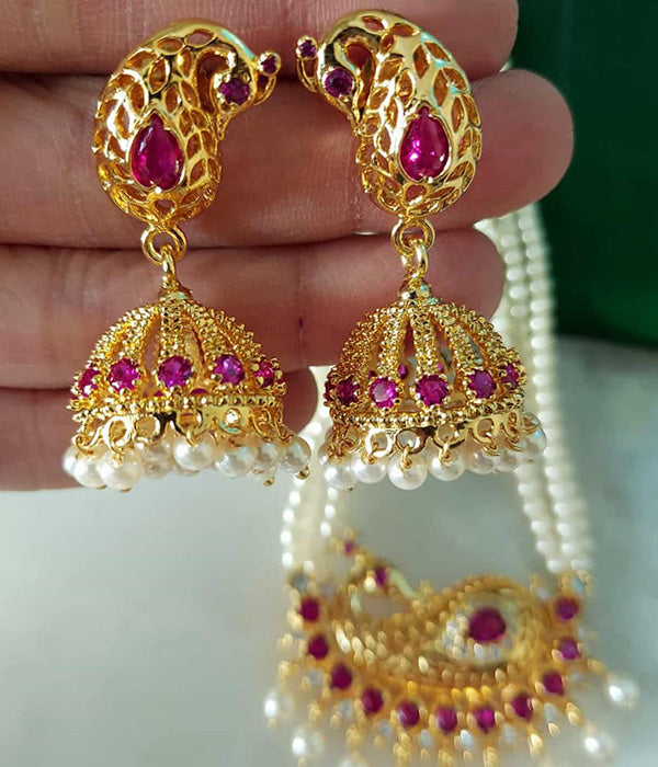 Elegant peacock-shaped gold-plated earrings with intricate ruby gemstone detailing and pearls hanging in a crescent-moon design, showcased on a hand against a green background.