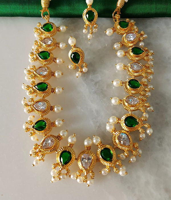 Ornate gold-toned necklace and earrings adorned with green gemstones and white pearls, displayed on a green textured background.