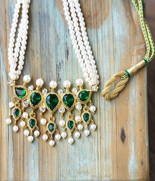Elegant green emerald and gold pendant necklace with pearls, placed on a rustic wooden background.