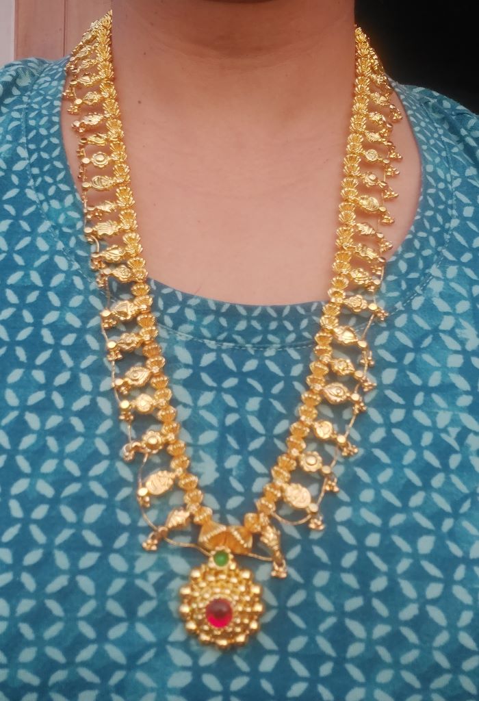 Golden Heavy Long Kolhapuri Saaj, a traditional Indian jewelry piece featuring a necklace with intricate golden designs and a central pendant, displayed against a patterned blue fabric background.
