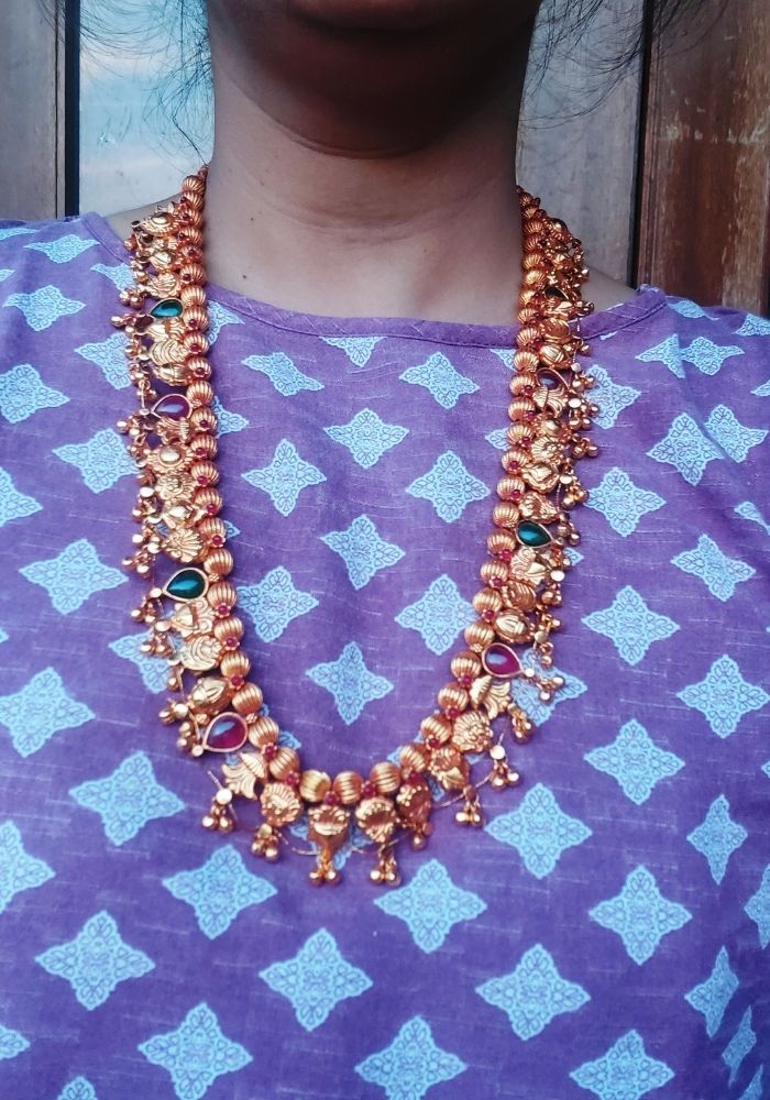 Geru Plated Kolhapuri Saaj, a traditional Indian necklace featuring intricate gold-toned beads and decorative patterns, placed against a vibrant purple background with a geometric star design.