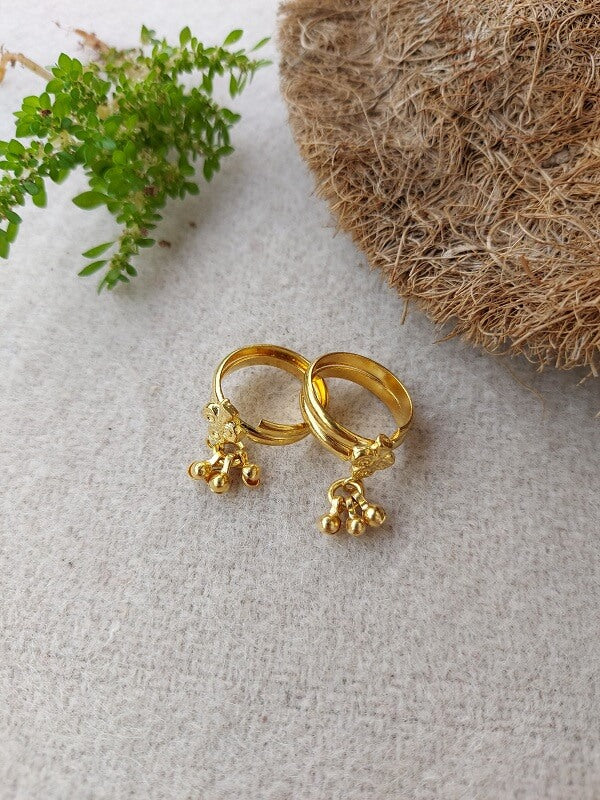 Elegant golden ghungroo toe rings with delicate jingling charms, set against a natural backdrop of green foliage and textured jute.