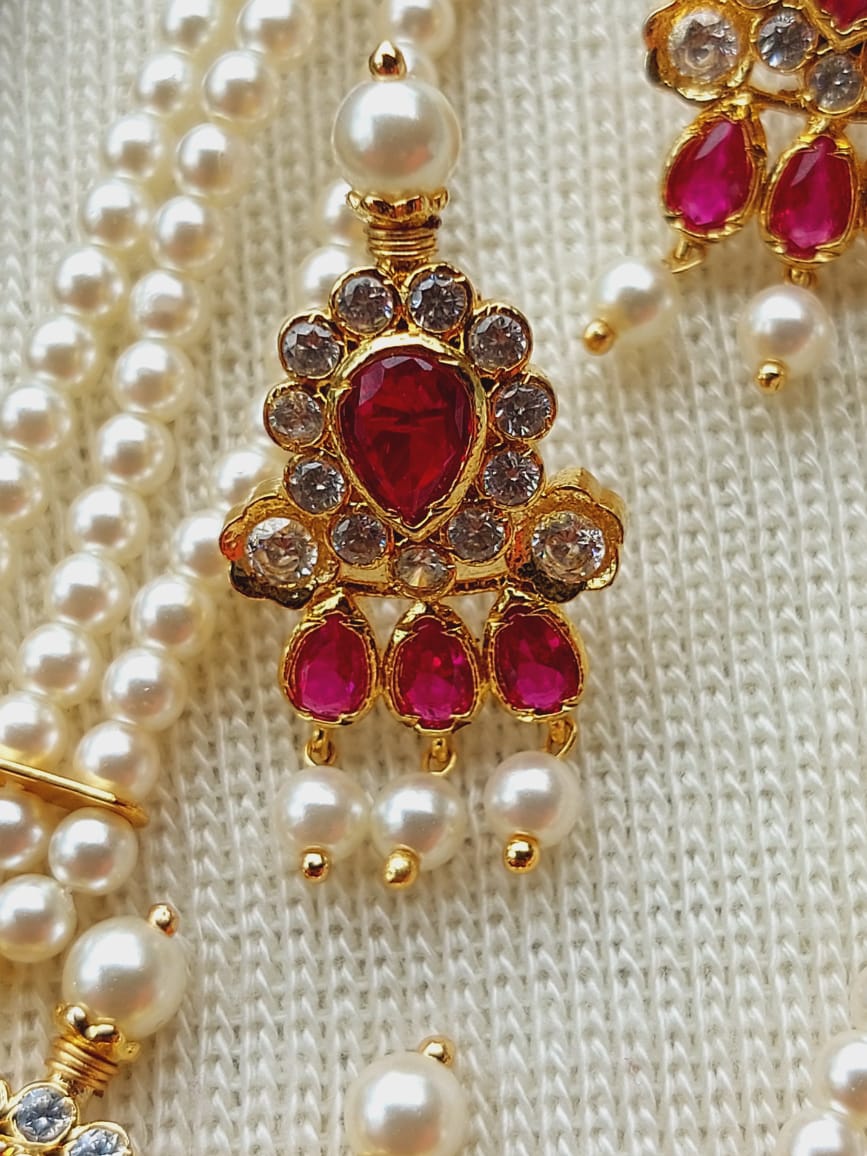 Ornate gold-toned necklace with a heart-shaped ruby centerpiece, surrounded by sparkling diamonds and dangling rubies, accented by strings of lustrous pearls on a textured white fabric background.