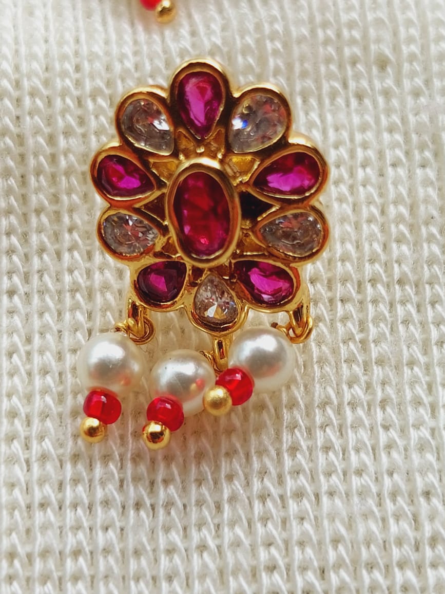 Elegant floral pendant with vibrant red and clear gemstones set in a gold-toned metal frame, accented with hanging pearl and red beads, displayed on a textured light-colored fabric surface.