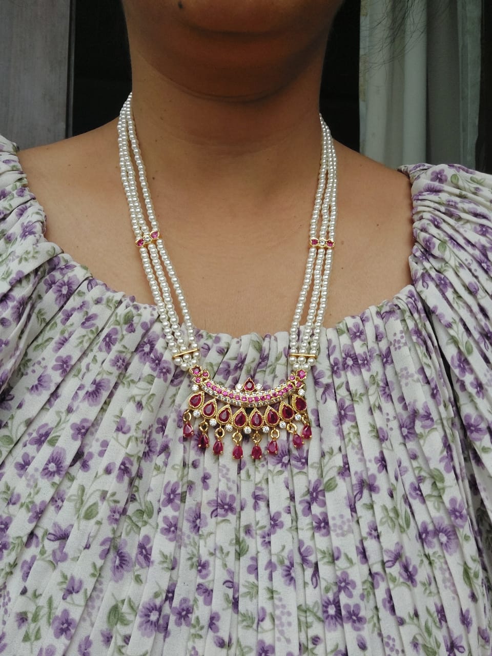 Designer Maharani Short Haar, multi-layered pearl necklace with red gemstone pendant, worn by a person in a floral print dress