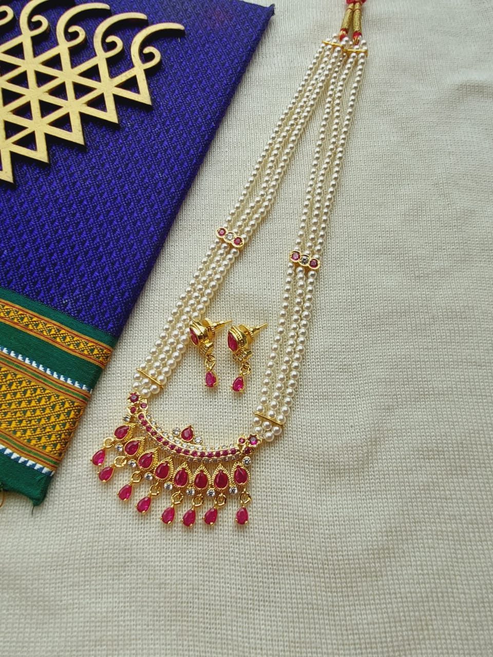 Elegant gold-toned Designer Maharani Short Haar necklace featuring intricate ruby-red gemstone accents, presented against a vibrant purple and green patterned textile background.